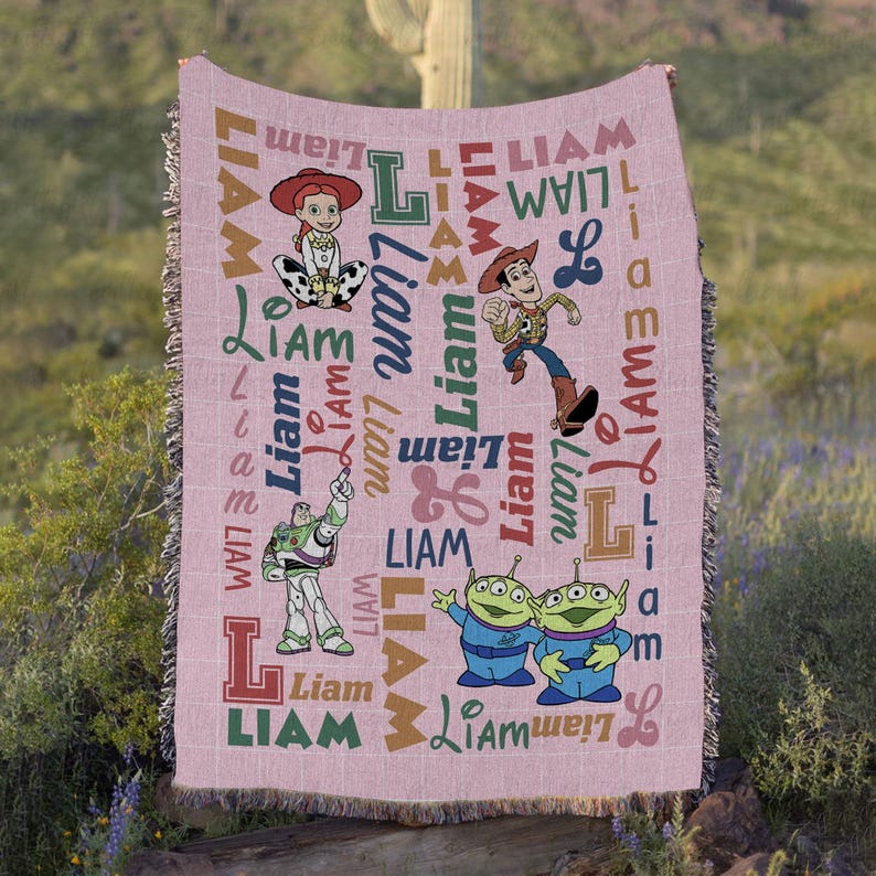 May include: A woven throw blanket in a light pink hue featuring a repeating pattern of the name Liam in various fonts and colors, alongside illustrations of Jessie, Woody, Buzz Lightyear, and two green aliens from the Toy Story film franchise.