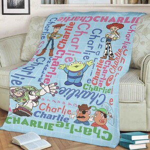 May include: A light blue fleece blanket featuring a grid pattern and the repeated name Charlie in various colors. The blanket displays Toy Story characters including Woody, Jessie, Buzz Lightyear, an Alien, and two Potato Head figures.