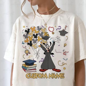 Puede incluir: Camiseta de color crema con un dise&ntilde;o tem&aacute;tico de graduaci&oacute;n. Presenta al personaje Goofy sosteniendo globos con forma de cabezas de Mickey Mouse. El dise&ntilde;o incluye birretes, libros y el texto "CUSTOM NAME".