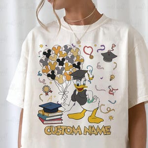 Puede incluir: Camiseta color crema con un dise&ntilde;o tem&aacute;tico de graduaci&oacute;n con el Pato Donald sosteniendo globos de Mickey Mouse. El dise&ntilde;o incluye gorras de graduaci&oacute;n, libros y el texto "CUSTOM NAME".