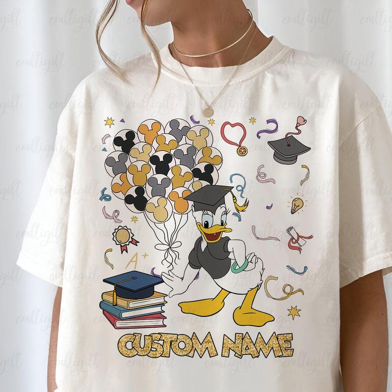 Puede incluir: Camiseta color crema con un dise&ntilde;o tem&aacute;tico de graduaci&oacute;n con el Pato Donald sosteniendo globos de Mickey Mouse. El dise&ntilde;o incluye gorras de graduaci&oacute;n, libros y el texto "CUSTOM NAME".