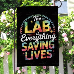 May include: Black garden flag with the text "The LAB is Everything SAVING LIVES" in a colorful tie-dye design. The flag is hanging on a black metal garden stake, with a white picket fence and pink flowers in the background.