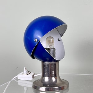 Könnte beinhalten: Vintage Schreibtischlampe in Blau und Silber. Die Lampe hat einen kugelförmigen, klappbaren blauen Schirm, der eine Glühbirne freilegt. Der Sockel ist silberfarben und die Lampe hat ein weißes Netzkabel. Retro-Design.