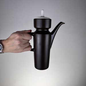Peut inclure: Une théière en céramique brune avec un couvercle blanc et un bec verseur. La théière a un design unique avec un corps rond et un col haut et fin.
