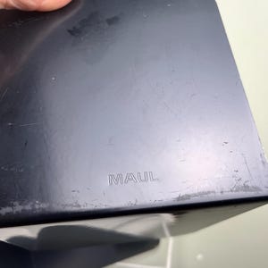 May include: A black, rectangular object with rounded corners. The word "MAUL" is embossed on the surface. The object appears to be made of a hard material, possibly plastic, and shows signs of wear.