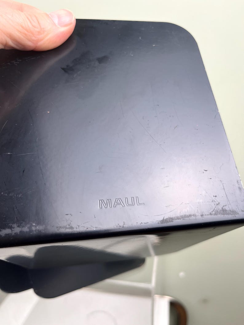 May include: A black, rectangular object with rounded corners. The word "MAUL" is embossed on the surface. The object appears to be made of a hard material, possibly plastic, and shows signs of wear.