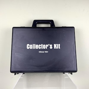 Könnte beinhalten: Ein schwarzer Sammlerkoffer mit Griff, mit dem Text "Collector's Kit" und dem Namen "Ottmar Hörl" in Weiß. Der rechteckige Koffer scheint aus Kunststoff zu sein.