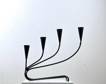 May include: A black metal candelabra with four candle holders. The candelabra has a curved design and is sitting on a white pedestal.