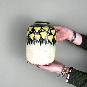 May include: A ceramic vase with a cream-colored base and a black, yellow, and green geometric design. The vase has a cylindrical shape and is held by hands.