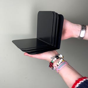 May include: A set of four black metal bookends. Each bookend has a right-angle design, with a flat base and a vertical support. The bookends are held in a person's hands.