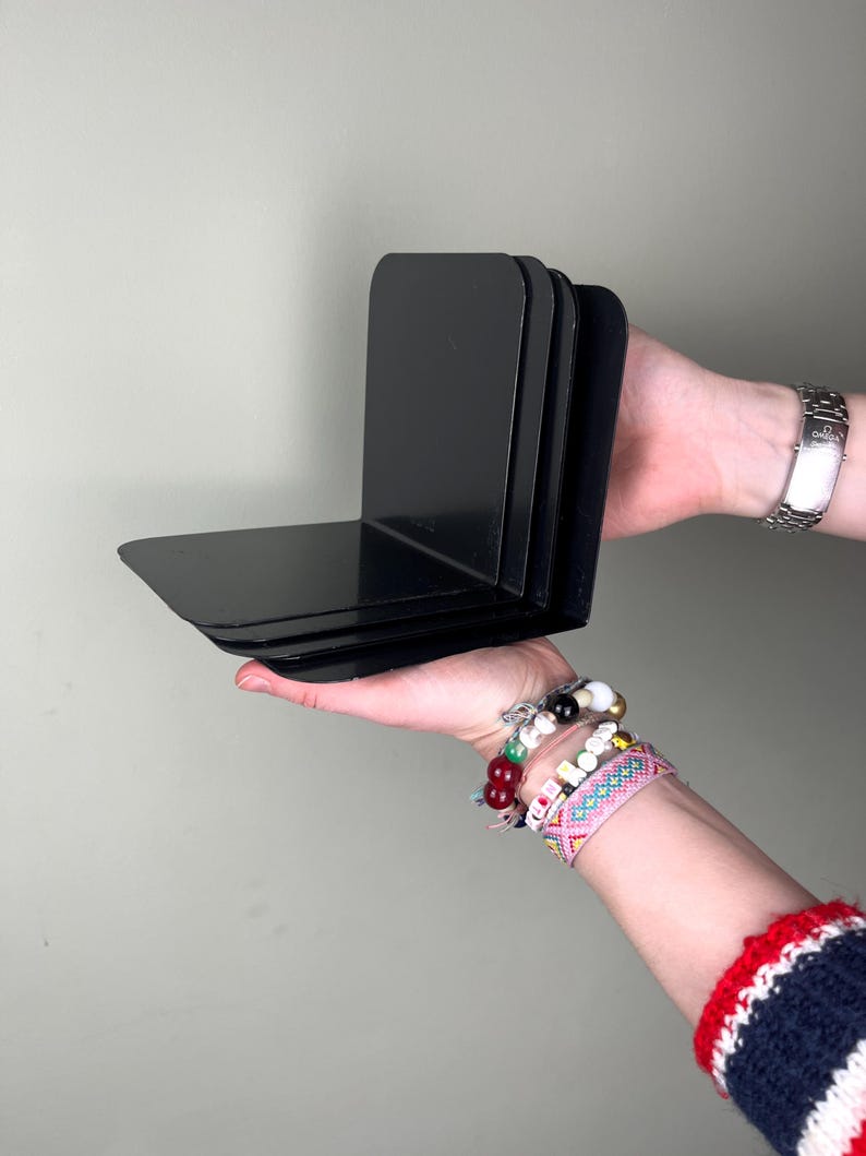 May include: A set of four black metal bookends. Each bookend has a right-angle design, with a flat base and a vertical support. The bookends are held in a person's hands.