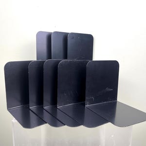 May include: A collection of six black metal bookends. Each bookend features a flat base and a vertical support with rounded corners. The bookends are arranged in a row, offering a functional and modern design for organising books on a shelf or desk.