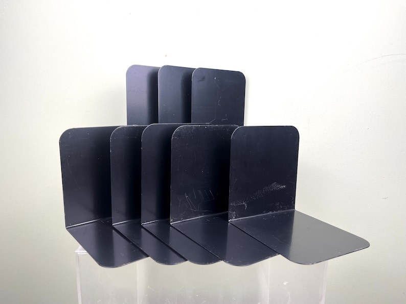 May include: A collection of six black metal bookends. Each bookend features a flat base and a vertical support with rounded corners. The bookends are arranged in a row, offering a functional and modern design for organising books on a shelf or desk.