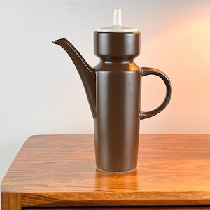 Peut inclure: Une cafetière en céramique brune avec un couvercle blanc et une poignée. La cafetière est posée sur une table en bois.
