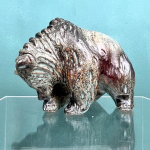 Puede incluir: Una escultura de cerámica de un oso con un esmalte marrón, verde y azul. El oso está de pie sobre una superficie de vidrio y mira hacia abajo.