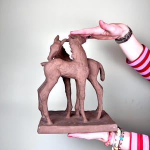 May include: A terracotta sculpture of two foals standing on a rectangular base. The foals are brown and have a textured surface.