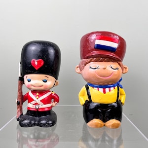 Può includere: Due statuine in ceramica di bambini in abiti tradizionali. Uno è un soldato con un cappello nero con un cuore rosso, una giacca rossa e stivali neri. L'altro è un ragazzo con un cappello rosso con una bandiera, una camicia gialla e una tuta nera.
