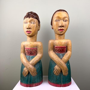 Könnte beinhalten: Zwei Holzstatuen von Figuren mit verschränkten Armen. Die Figuren haben helle Haut, rote Lippen und dunkles Haar. Sie tragen tealfarbene Kleider mit roten und goldenen Akzenten. Die Statuen werden auf einer weißen Oberfläche vor einer hellgrünen Wand ausgestellt.