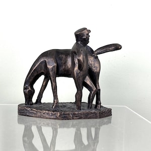 Könnte beinhalten: Bronzeskulptur, die einen Mann auf einem Pferd darstellt, mit einem zweiten Pferd in der Nähe. Die Skulptur hat eine dunkelbraune Patina und ist auf einem rechteckigen Sockel montiert. Das Pferd grast, und der Reiter trägt eine Kappe.