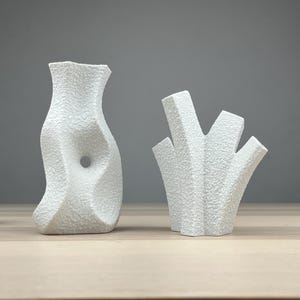 May include: Two white textured ceramic vases with unique shapes. One vase has an organic, curved form with a central hole. The other vase has a multi-stemmed, abstract design. A small wooden bird figurine is to the left.