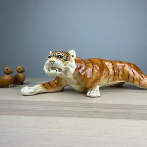 Może przedstawiać: Ceramiczna figurka tygrysa w pozycji kucznej, z pomarańczowymi i czarnymi paskami, białymi łapami i szczegółową twarzą. Dwie małe drewniane figurki ptaków znajdują się po lewej stronie tygrysa. Figurki znajdują się na jasnej powierzchni.