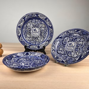 May include: Three round ceramic plates featuring a detailed blue and white floral and scrollwork pattern. Two plates are displayed upright on black stands, while the third plate lies flat in the foreground. The design includes intricate motifs.
