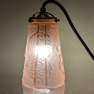 May include: A pink Art Deco style pendant light fixture with etched floral and geometric designs. The light fixture has a brass-colored top and a black cord. The light is on, illuminating the etched details.