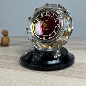 May include: A round table clock featuring a deep red face with gold-colored numerals and ornate hands, encased in a faceted clear glass housing. The clock is mounted on a black circular base with two silver-toned metal supports and a red second hand.