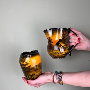 May include: Two ceramic vases with a brown and yellow glaze. Both vases feature a painted scene of a sunset over a countryside landscape. The vase on the left is smaller and has a rounded shape. The vase on the right is larger and has a more traditional pitcher shape with a handle.