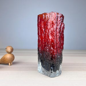 Peut inclure: Un vase cylindrique en verre présentant une surface texturée rappelant l'écorce. La couleur passe d'un noir profond à la base à un rouge translucide vibrant vers le haut, avec un bord inférieur en verre clair et un rebord irrégulier.