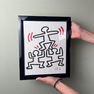 Könnte beinhalten: Schwarz gerahmtes Kunstwerk mit einer Illustration von Strichmännchen im Keith-Haring-Stil. Das Kunstwerk zeigt eine Pyramide von Figuren mit einer Figur an der Spitze, mit roten Linien. Das Kunstwerk ist signiert.