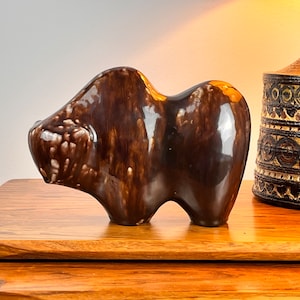 May include: A brown ceramic sculpture of a stylised animal, possibly a bear or a bull, with a glossy finish. The sculpture has a rounded, abstract form and is sitting on a wooden surface.