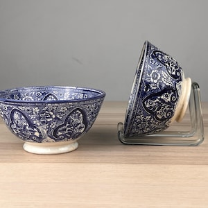 May include: Two ceramic bowls featuring a deep blue and white floral pattern. One bowl sits upright on its circular base, while the other is tilted on its side, supported by a clear acrylic stand, revealing the intricate botanical design.