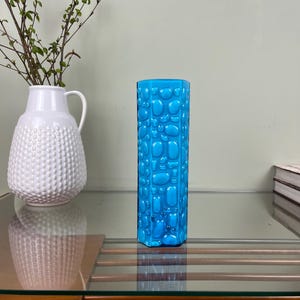 Puede incluir: Un jarrón turquesa cilíndrico con un patrón texturizado similar a guijarros. Junto a él, un jarrón de cerámica blanca con una superficie texturizada contiene ramas. Los objetos están sobre una mesa de cristal. Decoración del hogar.
