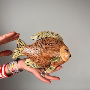 Könnte beinhalten: Eine Keramikfisch-Skulptur mit einem texturierten braunen Körper mit detaillierten Schuppen, einem glatten hellbeigen Gesicht und gelblichen Flossen mit dunkelbraunen Linien. Der Fisch hat ein kleines schwarzes Maul und ein dunkles Auge.