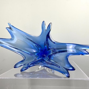 May include: A striking, star-shaped decorative bowl crafted from transparent blue glass. The bowl features a central, deep blue core with radiating, pointed edges. The glass has a glossy finish, reflecting light.