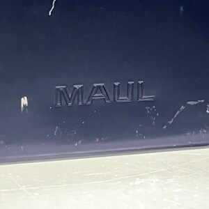 May include: A close-up shot of a dark blue surface with the embossed word "MAUL". The letters are raised and a darker shade of blue. The bottom of the image shows a lighter, possibly reflective surface.