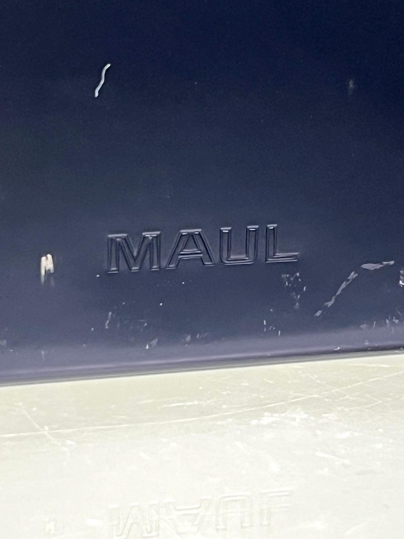 May include: A close-up shot of a dark blue surface with the embossed word "MAUL". The letters are raised and a darker shade of blue. The bottom of the image shows a lighter, possibly reflective surface.