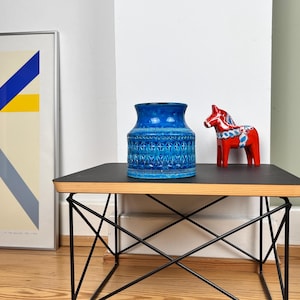 Könnte beinhalten: Eine blaue Keramikvase mit einem strukturierten geometrischen Muster am Körper und eine rote Pferdefigur mit weiß-blauen Bemalungen, beide auf einem schwarzen quadratischen Tisch mit dünner Holzkante und schwarzem Drahtgestell platziert.