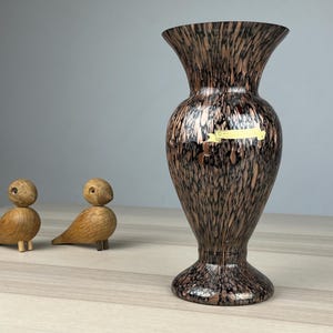 Könnte beinhalten: Eine hohe, gesprenkelte braun-schwarze Vase mit ausgestelltem Rand und breitem Fuß. Links von der Vase befinden sich zwei kleine Holzfiguren von Vögeln. Die Vase hat ein Etikett.