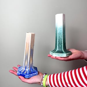 Könnte beinhalten: Zwei Keramikvasen mit sechseckigem Sockel. Die Vase links ist weiß mit blauen und goldenen Streifen. Die Vase rechts ist grün mit einem weißen Farbverlauf.