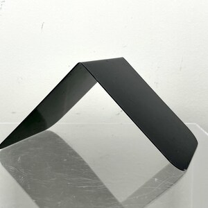May include: A black metal bookend with a minimalist, angled design. The bookend is bent at a 90-degree angle, forming a triangular shape. The surface is smooth and reflective, and it rests on a clear surface.