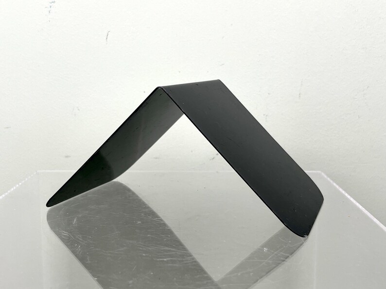 May include: A black metal bookend with a minimalist, angled design. The bookend is bent at a 90-degree angle, forming a triangular shape. The surface is smooth and reflective, and it rests on a clear surface.