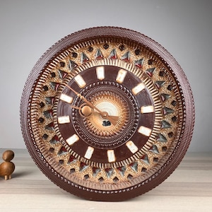 May include: A round ceramic wall clock featuring a dark brown outer rim with a textured pattern. The face displays a central sunburst design in cream and gold, surrounded by a dark brown ring with white rectangular hour markers and triangular accents.