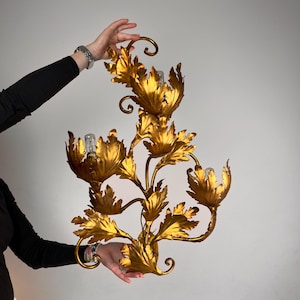 May include: A gold-coloured metal wall sconce featuring a floral design with three light bulb sockets. The fixture is shaped like a branching stem with multiple large, textured leaves and petals that curve outward in a decorative arrangement.