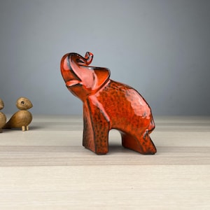 Könnte beinhalten: Eine glänzende, orange-braune Elefantenfigur aus Keramik. Links vom Elefanten befinden sich zwei kleine Vogelornamente aus Holz. Der Elefant ist das Hauptmerkmal dieser dekorativen Darstellung. Auf einer Holzoberfläche.