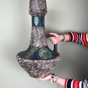 Op de afbeelding: Een grote, getextureerde keramische vaas met een donkere handgreep. De vaas heeft een gespikkelde, grijsbruine body met blauwe en groene accenten. Het ontwerp omvat een brede basis, een smalle hals en een uitlopende bovenkant. Een decoratief stuk voor in huis.