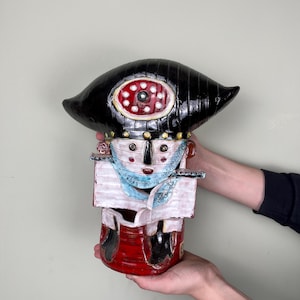 May include: A ceramic sculpture of a stylized figure with a large black hat, a white face, and a red, white, and blue patterned body. The sculpture is held in two hands.