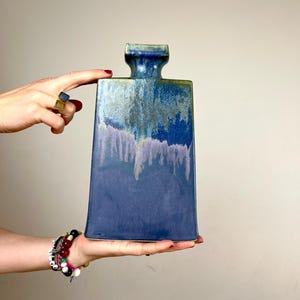 Könnte beinhalten: Eine rechteckige Keramikflasche mit engem Hals und ausgestelltem Rand. Die Oberfläche ist mit einer reaktiven Glasur in tiefen Blau-, Indigo- und gedämpften Lavendeltönen versehen, die einen tropfenden, geschichteten Effekt aufweist.