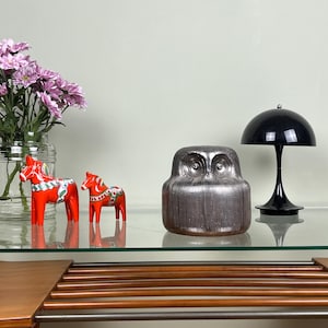 Könnte beinhalten: Eine klare Glasvase mit rosa Chrysanthemen, zwei orangefarbene Dala-Pferde mit bemalten Details, eine metallisch silberne Eulenfigur mit strukturierter Oberfläche und eine schwarze, kuppelförmige Tischlampe mit schlankem Fuß.